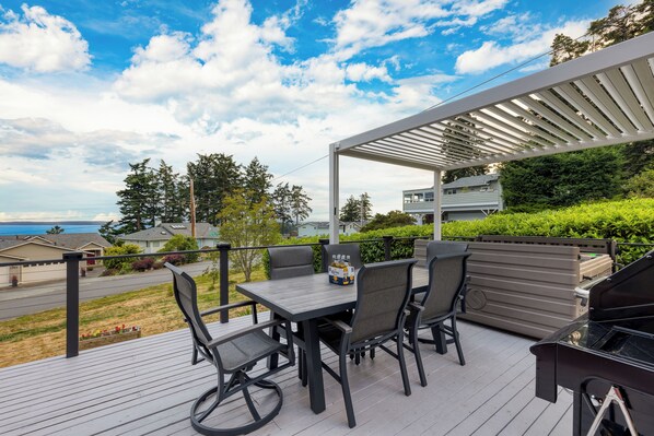 A deck with a table and chairs, a grill, and a view of the ocean and houses.