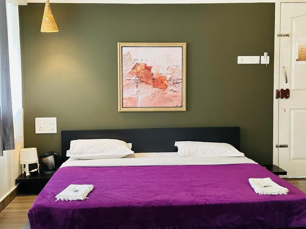 A hotel room with a bed, a purple bedspread, a framed artwork on the wall, a nightstand with a lamp, and a door with a handle.