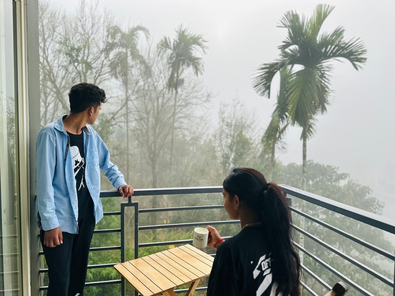 Two people on a balcony with a table and chairs, overlooking palm trees and a foggy landscape.