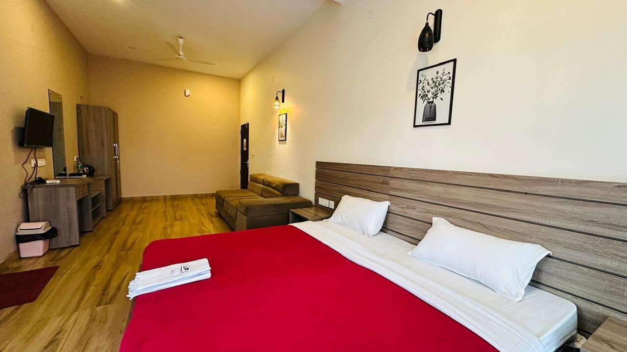 A hotel room with a wooden floor, a bed with white bedding, a red blanket, a wooden headboard, a TV on a stand, a wardrobe, and a small table with a lamp.