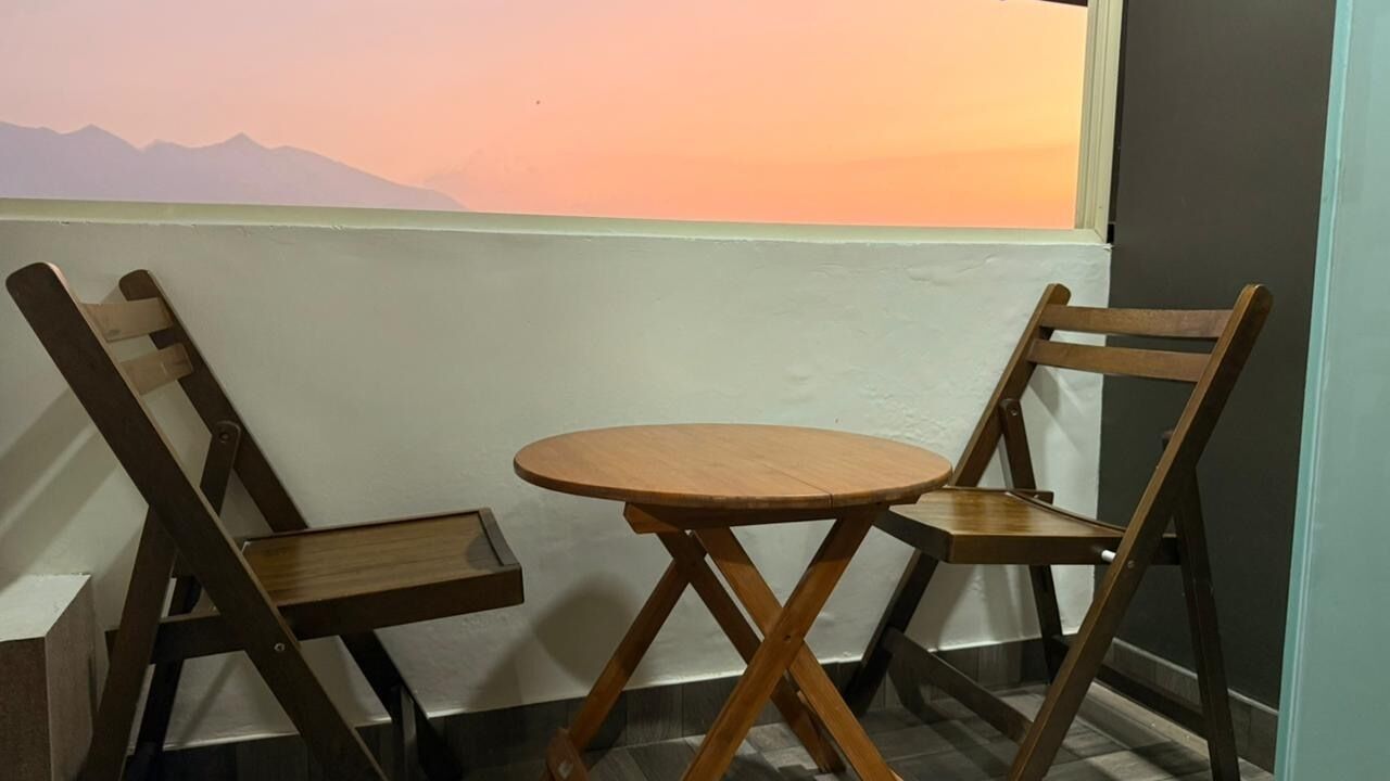 Een hoek met twee houten stoelen en een kleine ronde tafel, een raam met uitzicht op bergen, en een eenvoudige muur.