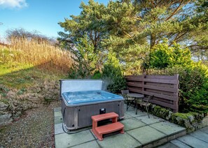 Un jacuzzi con un banco de madera y una pequeña mesa, rodeado de follaje y un muro de piedra.