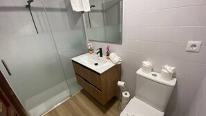 Un baño moderno con cabina de ducha de vidrio, tocador de madera, lavamanos blanco, espejo y retrete.
