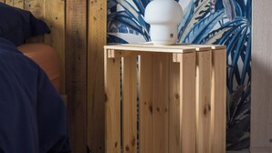 Ein Zimmer mit einem hölzernen Bett, einer blauen Decke, einer weißen Lampe auf einer Holzkiste und einer Wand mit tropischem Blattmuster.