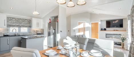 A modern kitchen with a built-in island, a dining area with a wooden table set for four, and a living area with a sofa and TV.