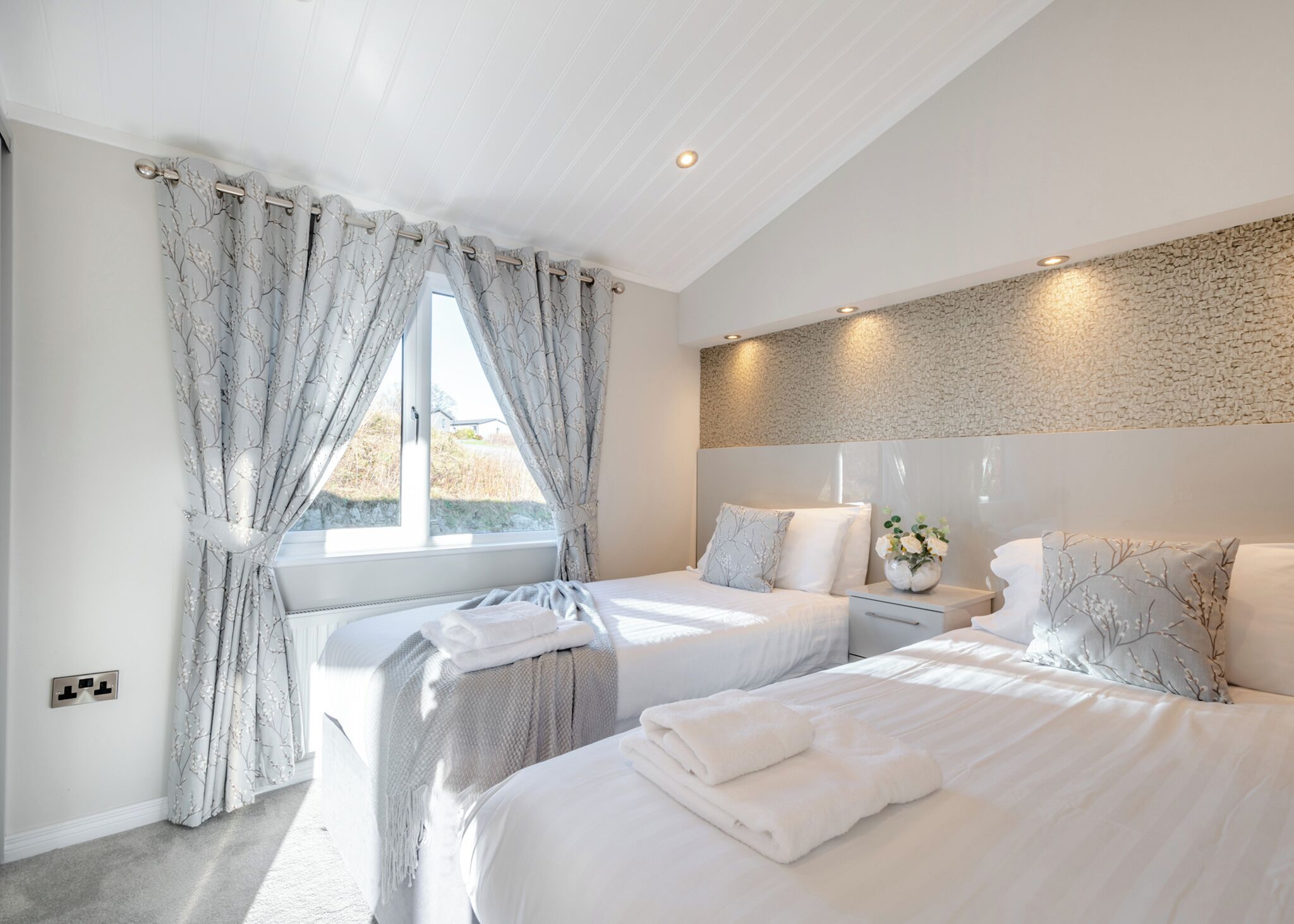 A modern bedroom with two beds, a large window with curtains, a stone-textured wall, and a bedside table with a vase.