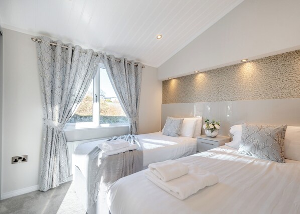 A modern bedroom with two beds, a large window with curtains, a stone-textured wall, and a bedside table with a vase. - Nith Valley Lodge Pet (Dumfries, Dumfries & Galloway)