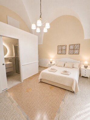 Een slaapkamer met een groot bed, twee nachtkastjes, een kroonluchter en een badkamer met een spiegel en wastafel.