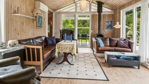 A cozy room with wooden walls and ceiling, a fireplace, two leather sofas, a small table with a floral tablecloth, and a view of a garden through a glass door.