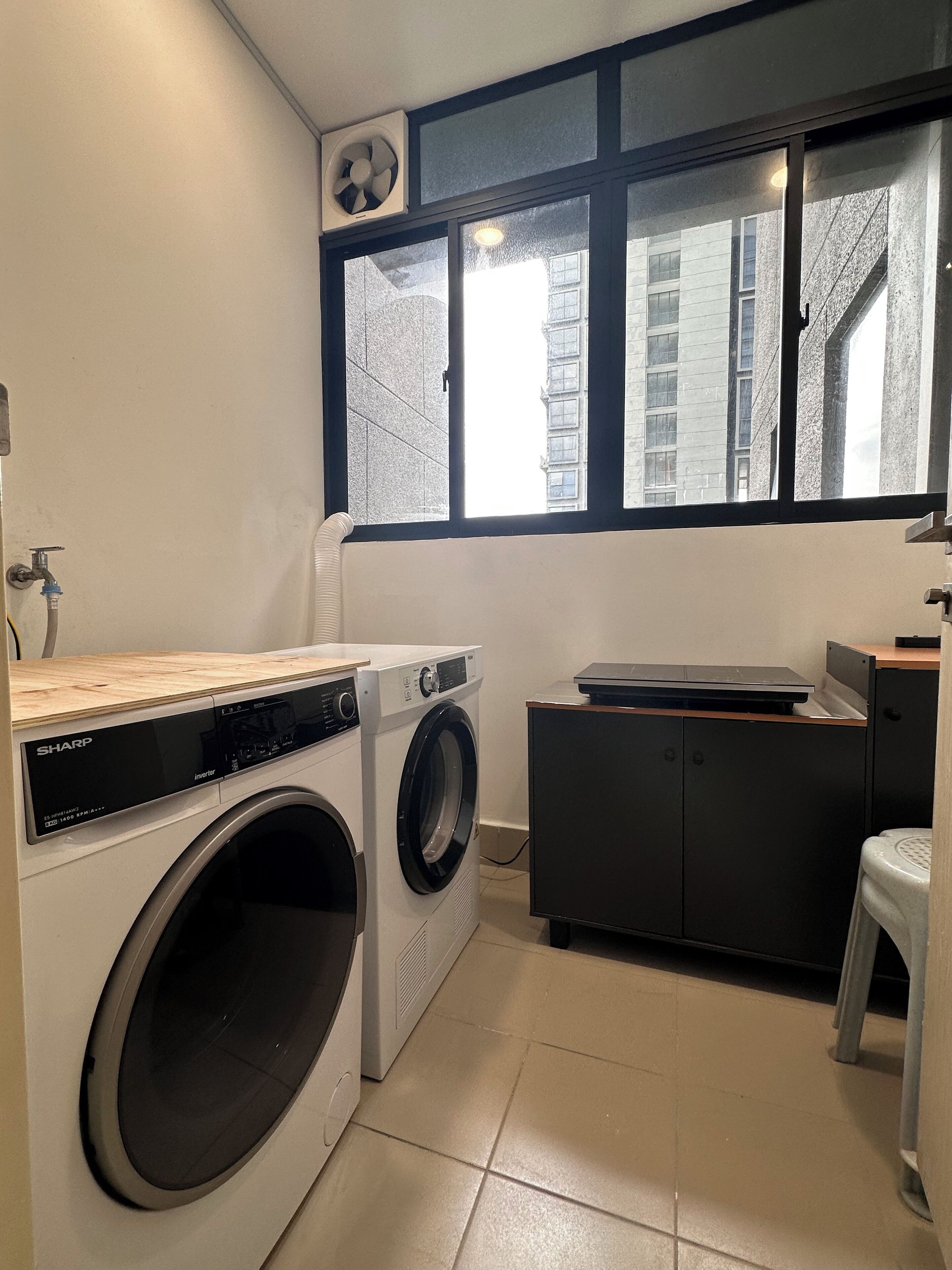A compact laundry room with a front-loading washing machine, a dryer, a small cabinet, and a window with a view of buildings outside.