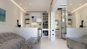 Ein modernes Wohnzimmer mit einem Glastisch, einem grauen Sofa und einer Küchenzeile mit weißen Schränken und Geräten.