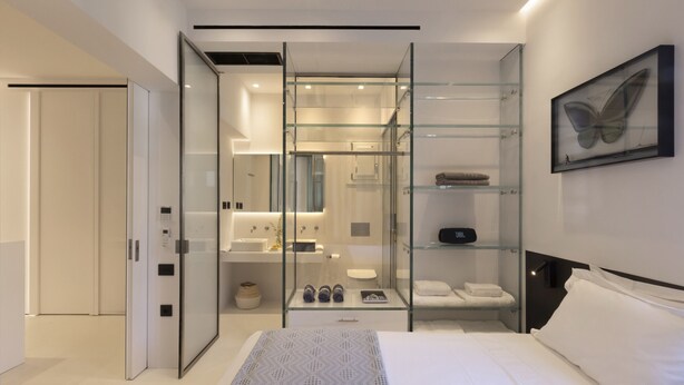 Ein modernes Hotelzimmer mit einer gläsernen Dusche, einem Bett mit gemustertem Überwurf, einem Wandbild mit Schmetterlingen und einem JBL-Lautsprecher auf einem Regal.