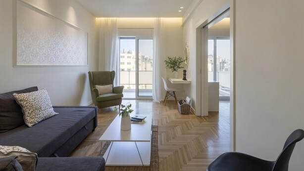 A modern living room with a sofa, armchair, and coffee table. There is a view of the city through large windows.