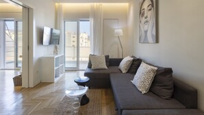 A modern living room with a grey sofa, a flat-screen TV, a floor lamp, and a large window with curtains.