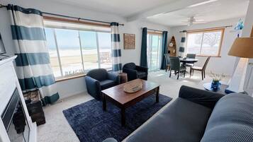 Un salon comprenant un canapé, des fauteuils, une table basse et un coin repas avec des chaises et une table. De grandes fenêtres sont présentes, avec des rideaux, un ventilateur de plafond et une vue sur la plage.