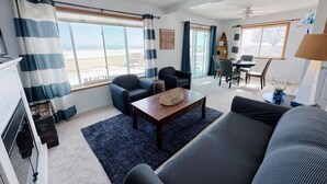 Ein Wohnzimmer mit Sofa, Sesseln, Couchtisch und Essbereich mit Stühlen und Tisch. Große Fenster mit Vorhängen, ein Deckenventilator und Blick auf den Strand.
