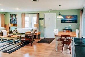 A living room with a sofa, armchairs, a coffee table, and a dining table with chairs. There is a painting on the wall, a lamp, and a door labeled "LAKE".