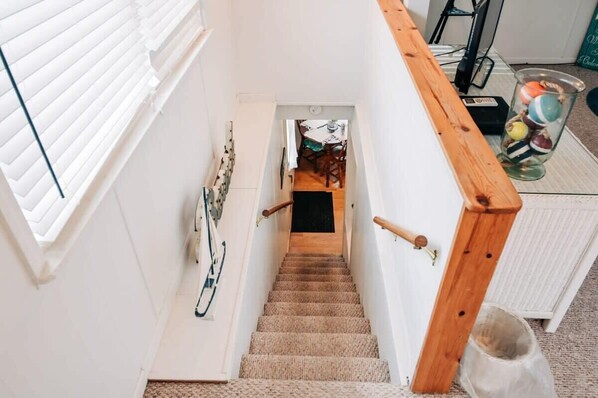 A staircase with a wooden handrail, a white wall, and a door leading to a room with a table and chairs.