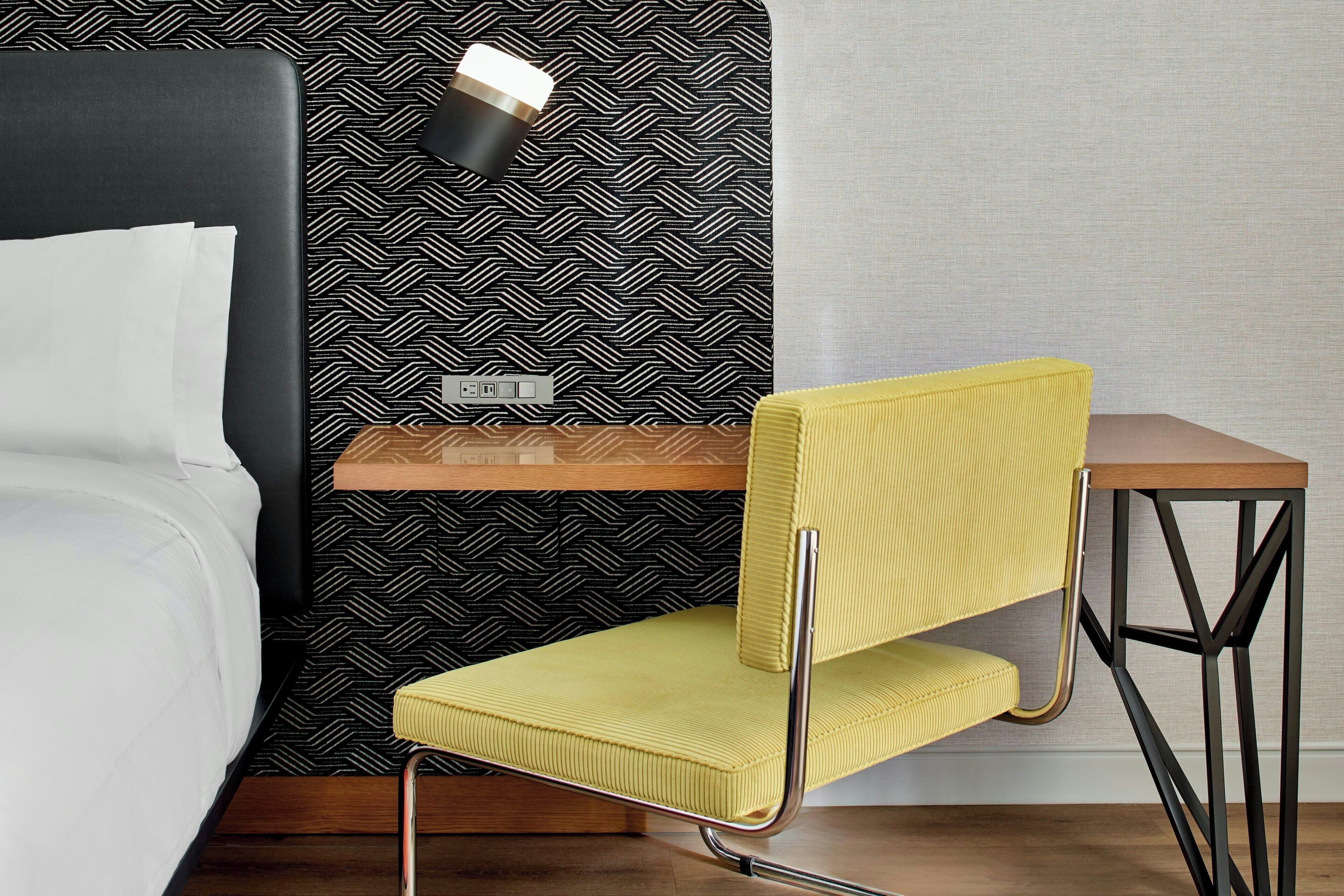 Una habitación de hotel moderna con una cama, un sillón amarillo, una mesita de madera y una lámpara de pared.