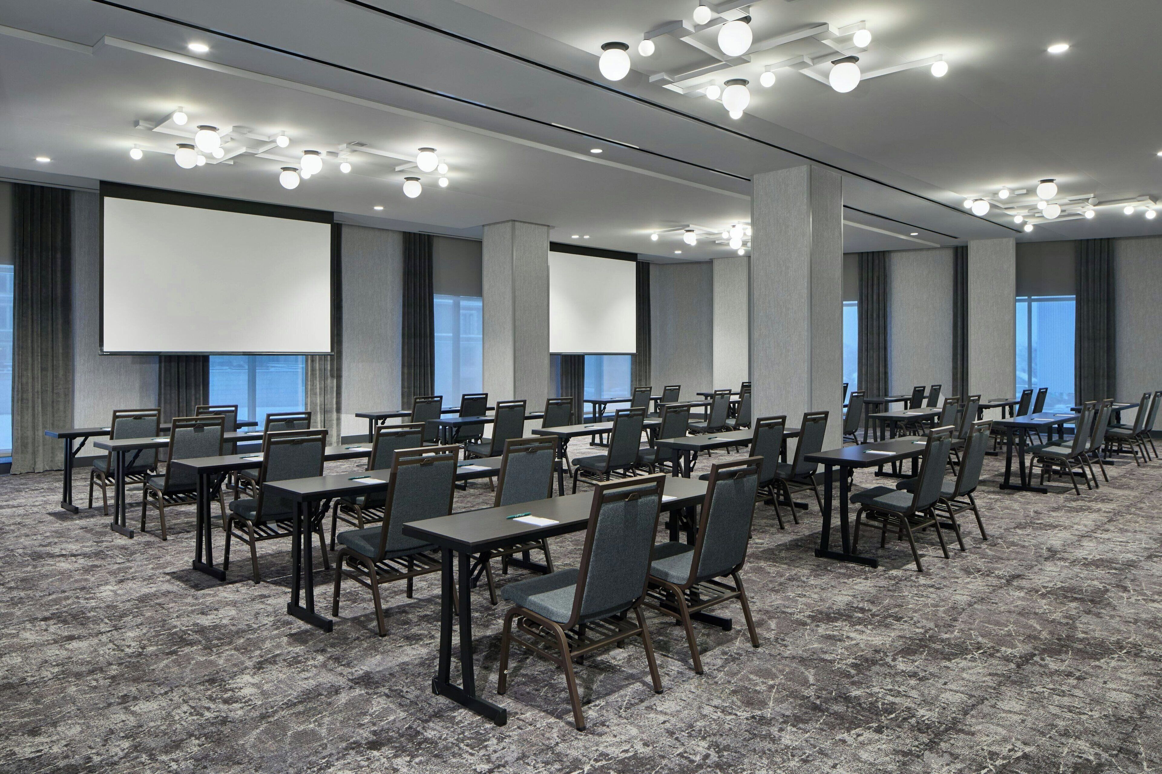 A conference room with rows of tables and chairs, large screens, and modern lighting fixtures.