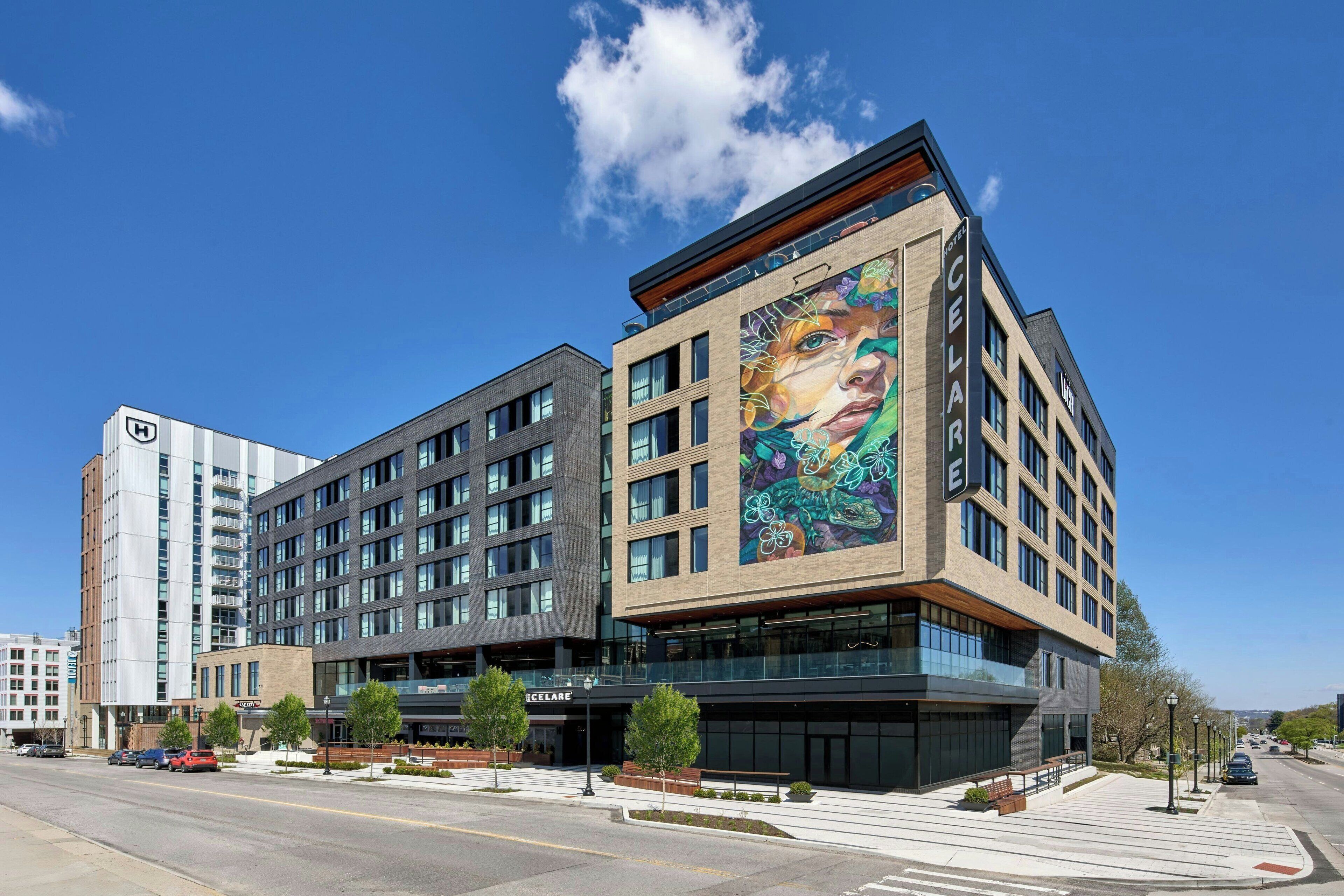 A modern hotel with a large mural on the facade, multiple floors, and a parking lot in front.