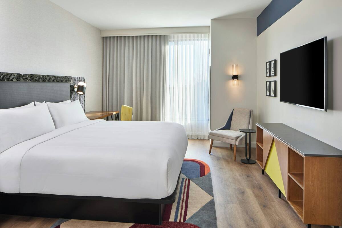 A hotel room with a large bed, a TV mounted on the wall, a desk with a chair, and a window with curtains.