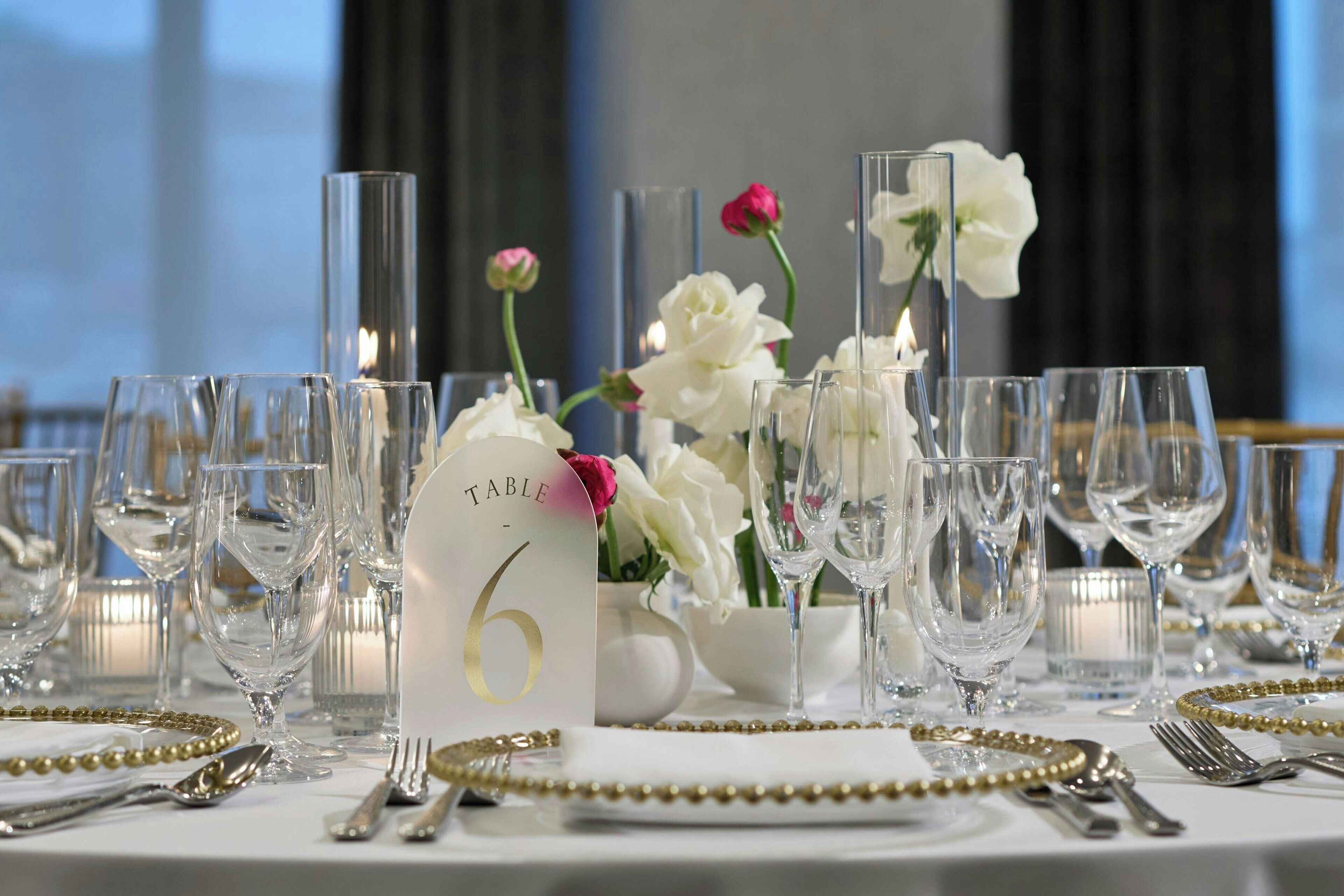 Mesa dispuesta para un evento formal, con sillas numeradas, cristalería transparente y flores decorativas.