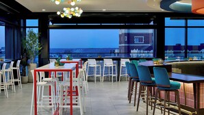 A modern bar area with white and teal chairs, a red-framed table, and a view of a cityscape at dusk. - Hotel Celare, Cincinnati Midtown, a Tribute Portfolio Hotel (Cincinnati)