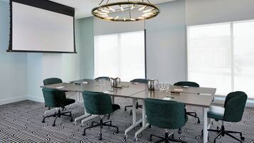 Un salón de conferencias con una amplia mesa, sillas verdes, una pantalla de proyector y una lámpara de araña moderna.