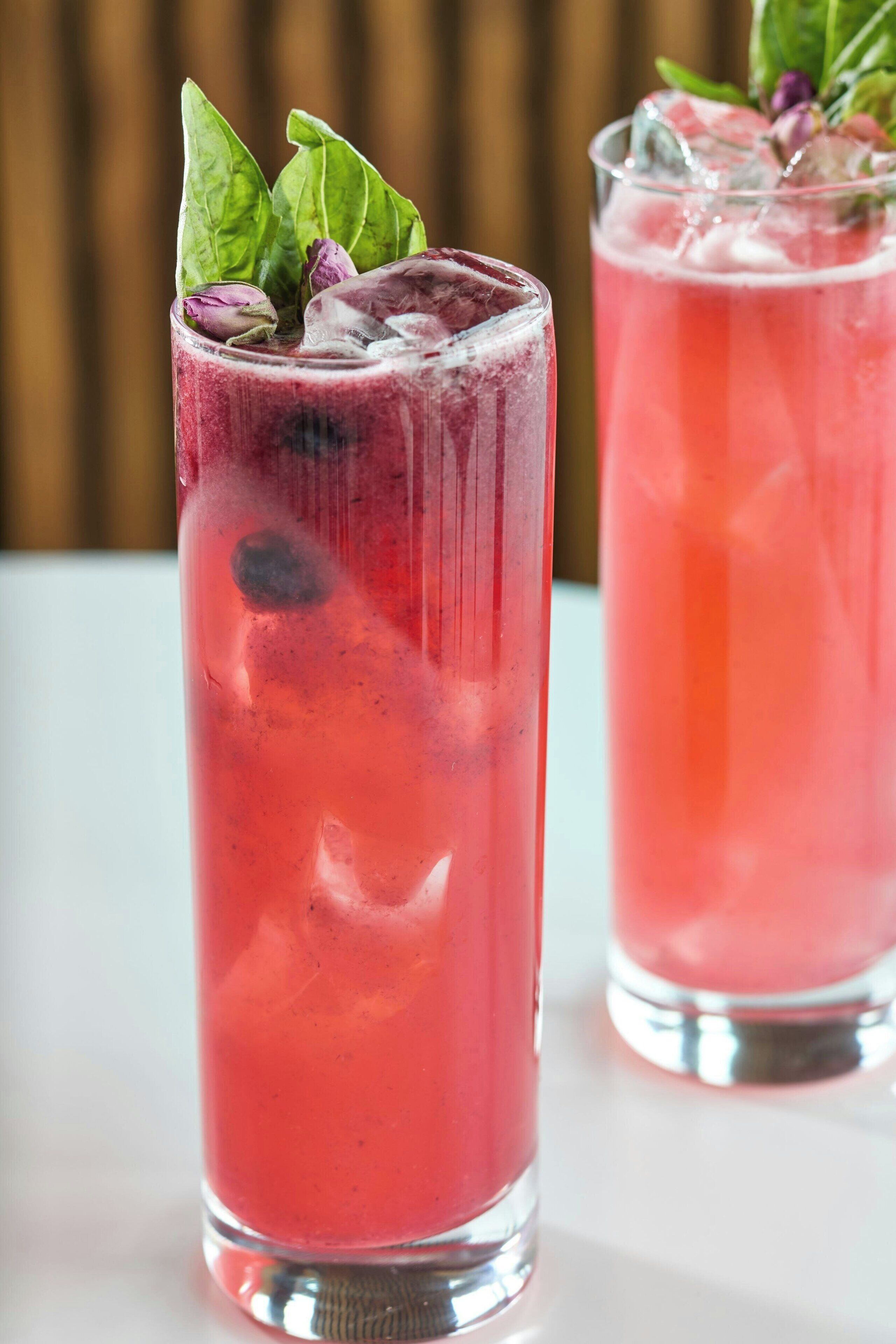 Two tall glasses of red beverage with ice and garnishes, placed on a white surface.