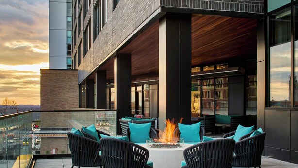 A modern outdoor seating area with a fire feature, surrounded by black wicker chairs and teal cushions, overlooking a cityscape at sunset. - Hotel Celare, Cincinnati Midtown, a Tribute Portfolio Hotel (Cincinnati)