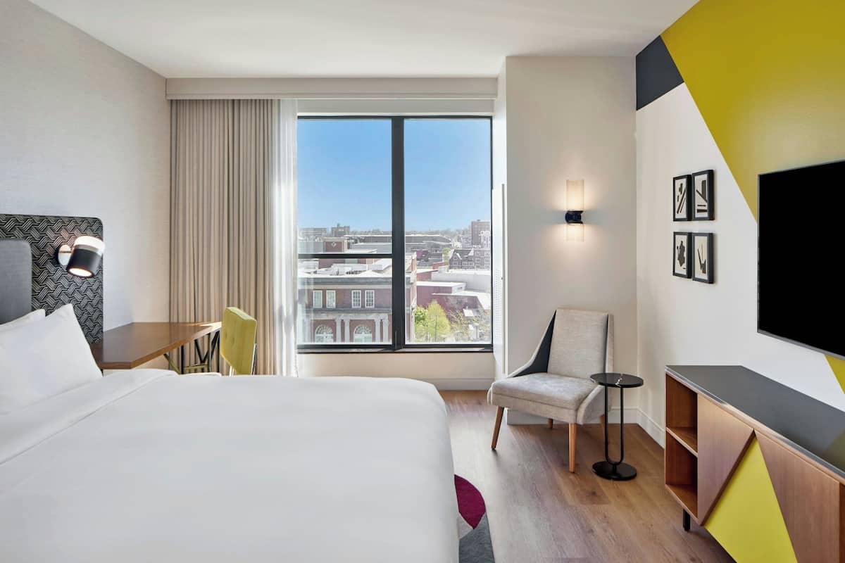 A hotel room with a large bed, a desk, a chair, a TV, and a window with a city view.