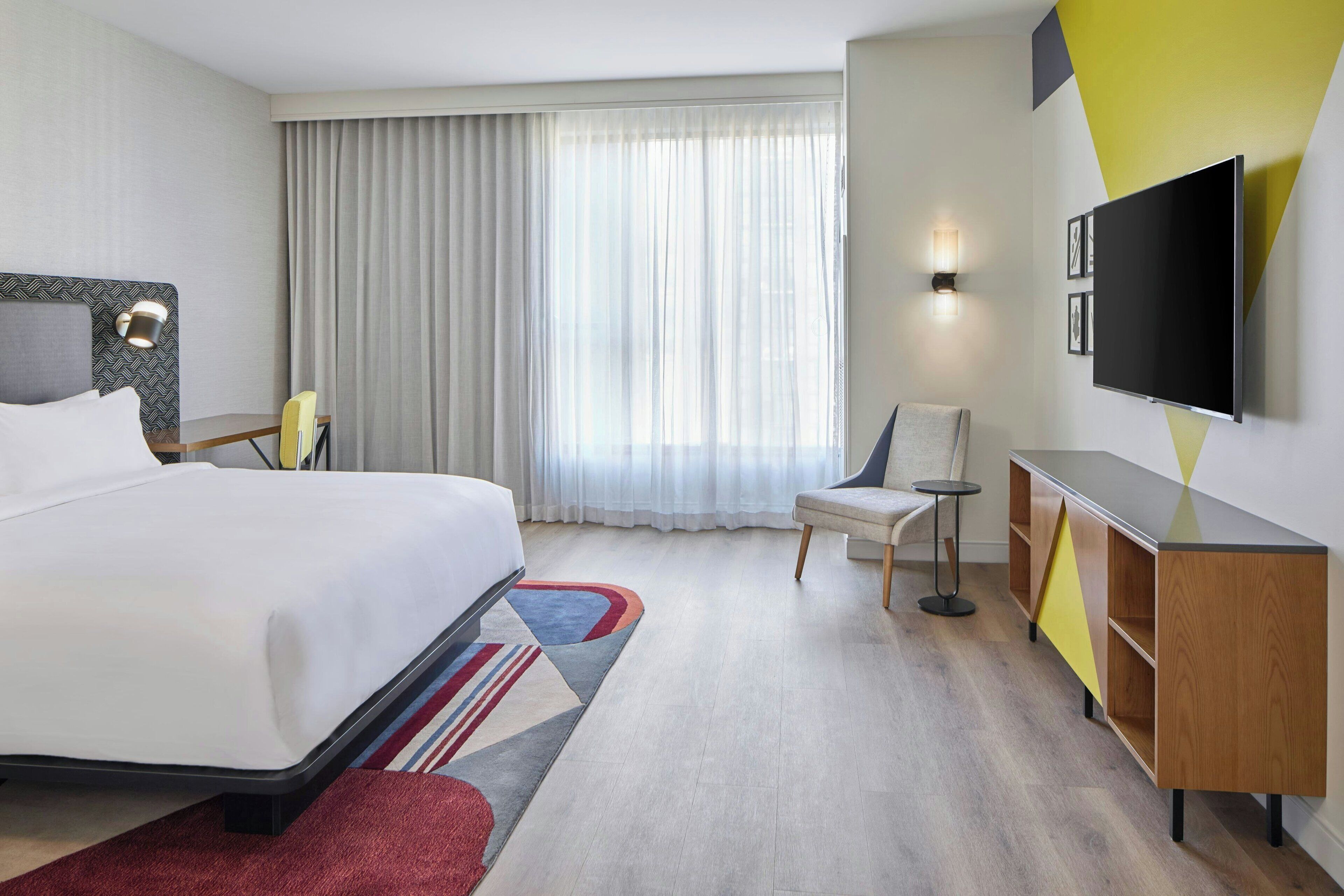 A hotel room with a bed, a desk, a chair, a TV, and a window with curtains.