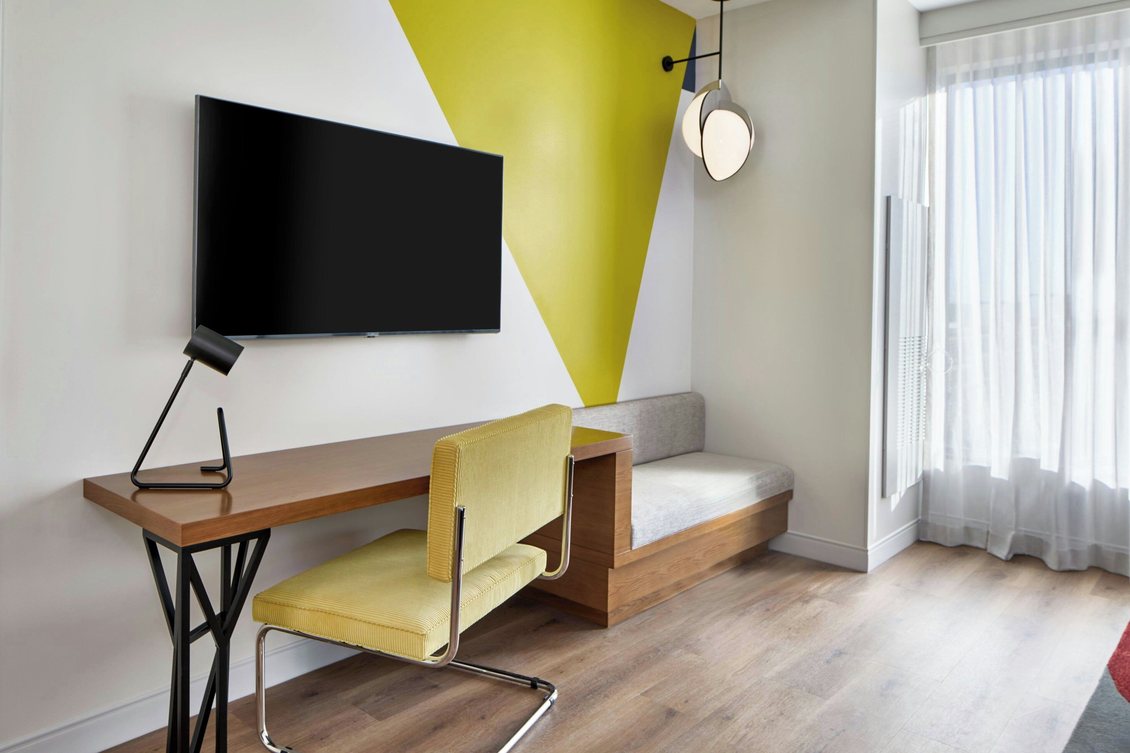 A modern room with a wooden desk, a yellow chair, a mounted TV, and a window with sheer curtains.