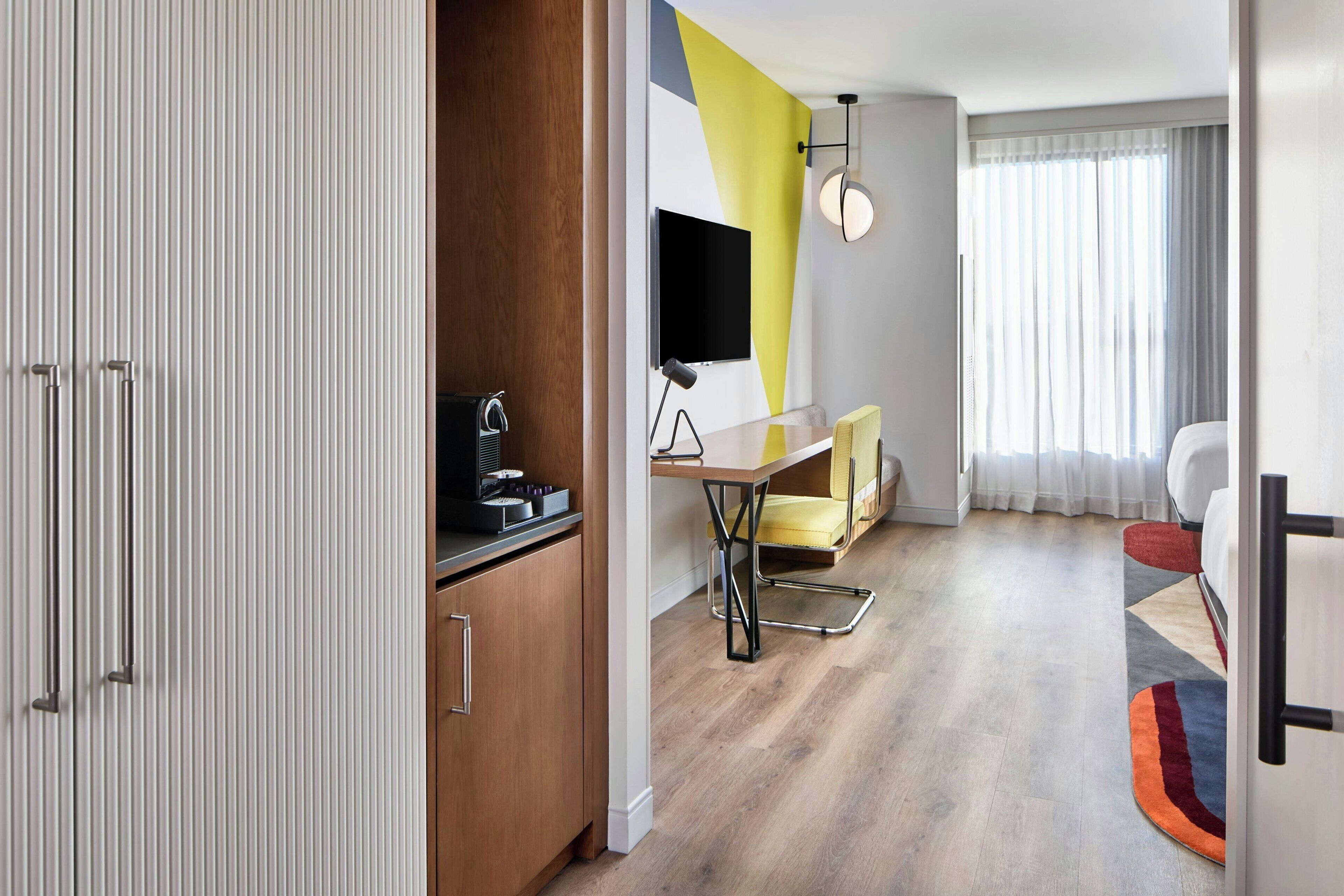 A modern hotel room with a wooden wardrobe, a desk with a chair, a flat-screen TV, and a large window with curtains.