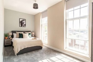A bedroom with a bed, bedside table, lamp, and a large window with a view of buildings.