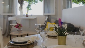 Un comedor con una mesa lista para dos personas, una planta en maceta y vistas a árboles a través de ventanas abiertas.