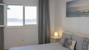 Un dormitorio con cama, mesita de noche, lámpara, ventana con vista al mar y un cuadro en la pared.