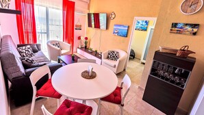 Una sala de estar moderna con una mesa redonda blanca, sillas con cojines rojos, un sofá gris y una pequeña zona de cocina con un soporte para vinos.