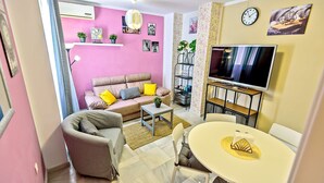 A compact living space with a pink wall, a sofa with cushions, a coffee table, a TV on a stand, and a small bookshelf.