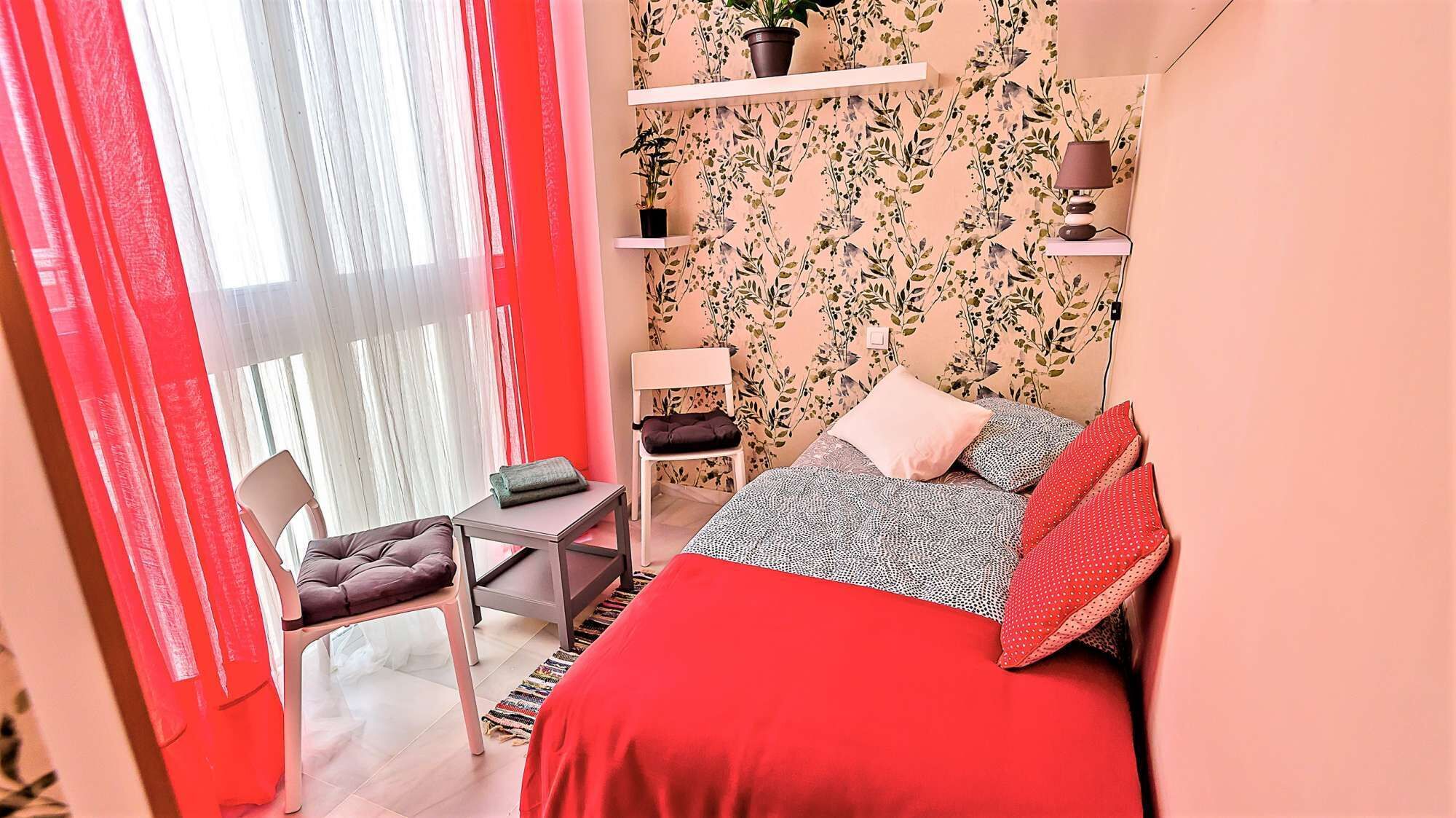 Une chambre à coucher de taille réduite, comprenant un lit, une chaise, une petite table, une étagère avec des plantes et une lampe.