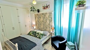 A bedroom with a bed, a chair, a wardrobe, a bedside table with lamps, and a window with curtains.