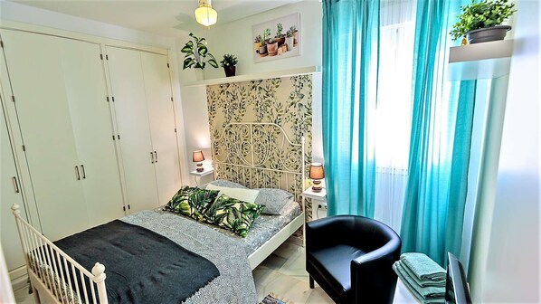 A bedroom with a bed, a chair, a wardrobe, a bedside table with lamps, and a window with curtains.