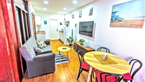 A compact living space with a sofa, a dining table with chairs, a TV mounted on the wall, and a small kitchen area.