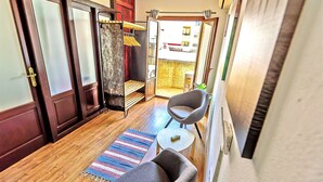 A hallway with a wooden floor, a striped rug, a grey armchair, a small round table with a remote control, and a door with a towel rack.