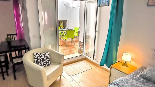 A living room with a sofa, a small table with a lamp, and a sliding glass door leading to a dining area with chairs and a table.