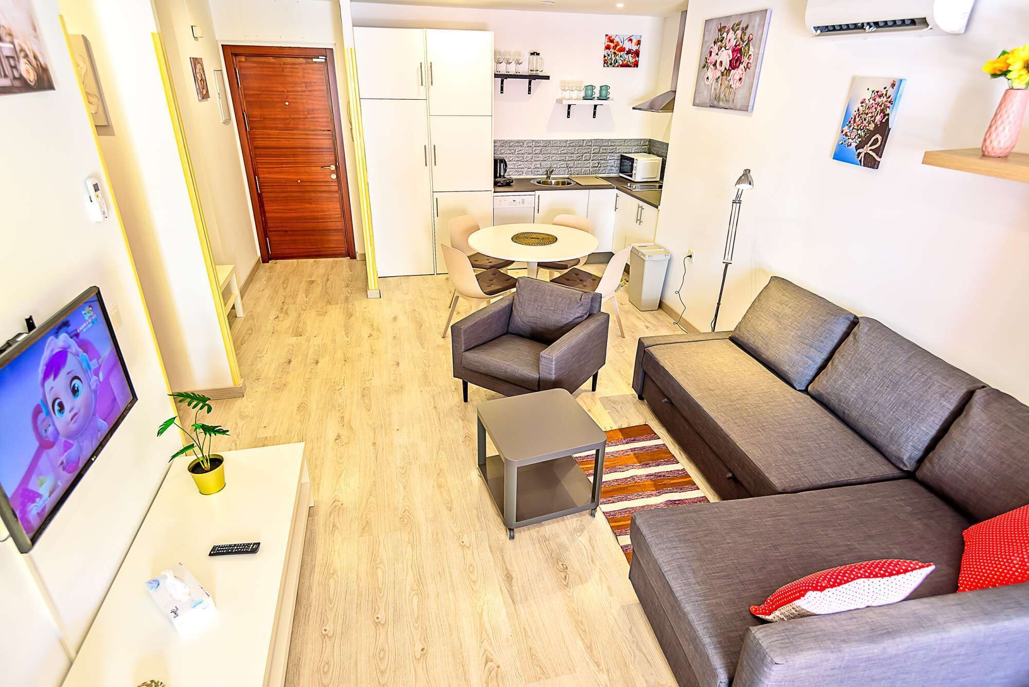 A modern living room with a grey sofa, a small coffee table, and a TV. There is a kitchen area with a dining table and chairs.