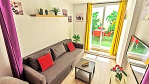 A living room with a sofa, a small table, and a TV. There are plants on the windowsill and a yellow curtain. - Plaza Herrería Central Apartment "D" with Terrace. Right downtown, best location (El Puerto de Santa María)