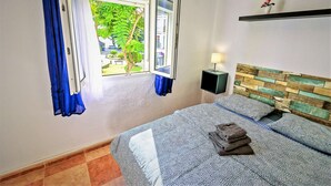 A bedroom with a bed, a nightstand, a window with curtains, and a view of greenery outside. - Plaza Polvorista 4-Bedroom Apartment with Balcony in front of the beautiful park (El Puerto de Santa María)
