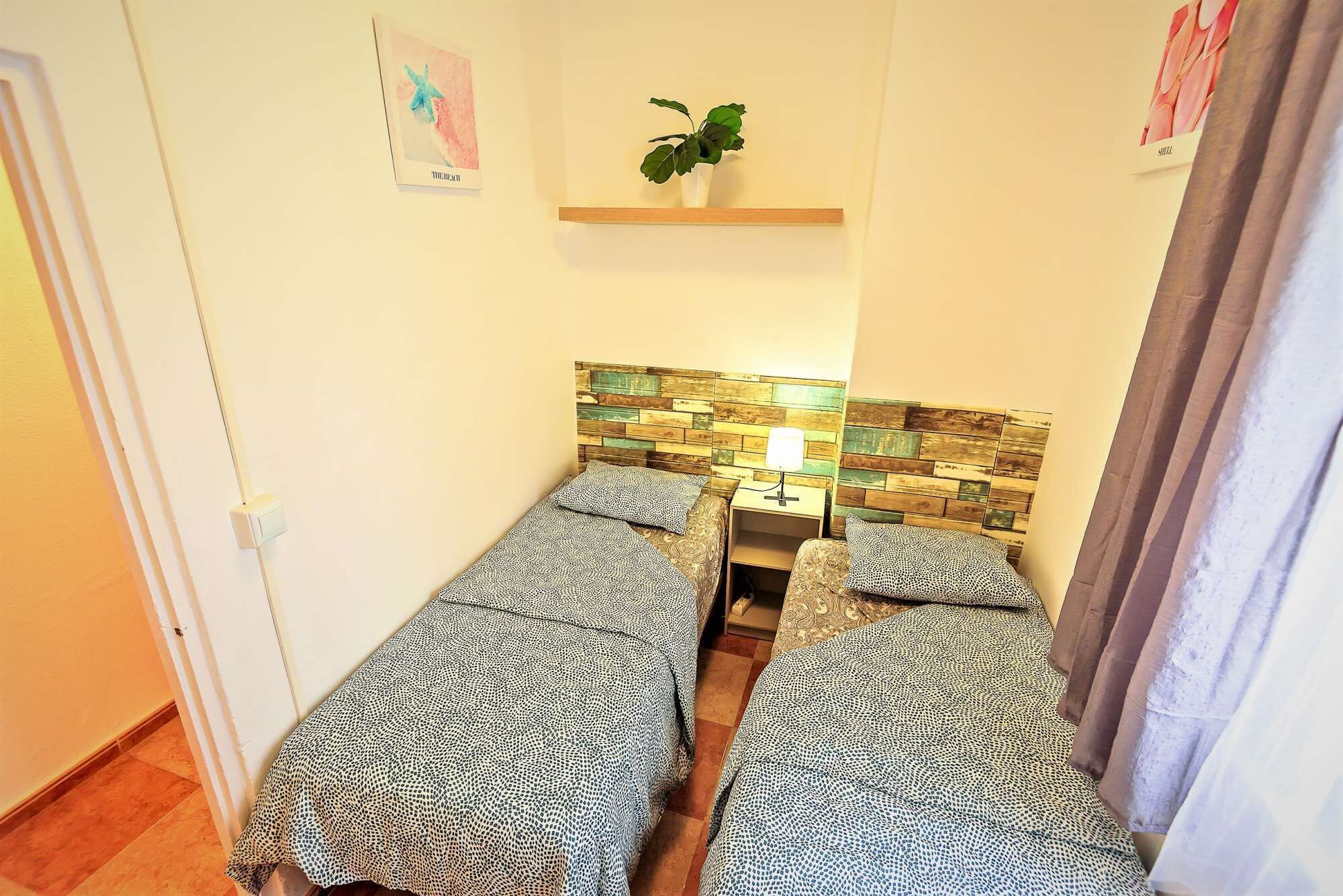A small, cozy room with two beds, a nightstand, a lamp, and a plant.